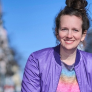 Foto von Kati Bohnet, lächelnd, in der Straße einer Großstadt. Blauer Himmel.