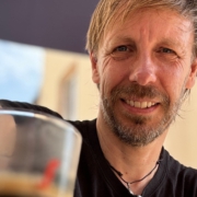 Foto von Christopher End, lächelnd. Im Hintergrund ein Sonnenschirm, vor ihmauf dem Tisch ein Espresso. Urlaubsstimmung.