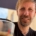 Foto von Christopher End, lächelnd. Im Hintergrund ein Sonnenschirm, vor ihmauf dem Tisch ein Espresso. Urlaubsstimmung.