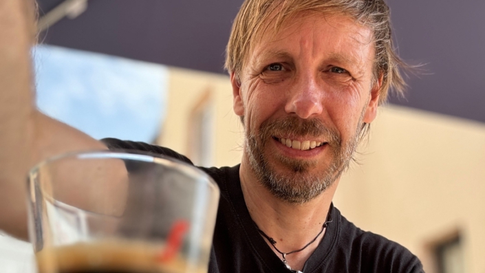 Foto von Christopher End, lächelnd. Im Hintergrund ein Sonnenschirm, vor ihmauf dem Tisch ein Espresso. Urlaubsstimmung.