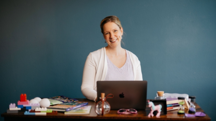 Foto von Diana Wegel, lächelnd am Schreibtisch mit Laptop und Lernutensilien