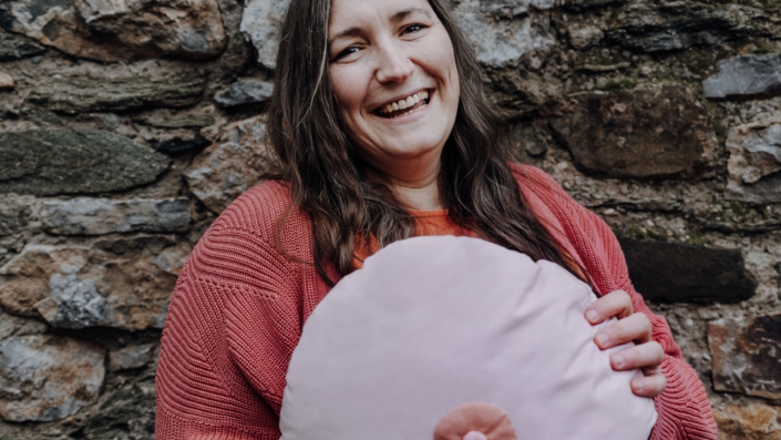 Porträtfoto von Ramona Noll, die ein große Kissen in Brustform vor sic hält und lacht